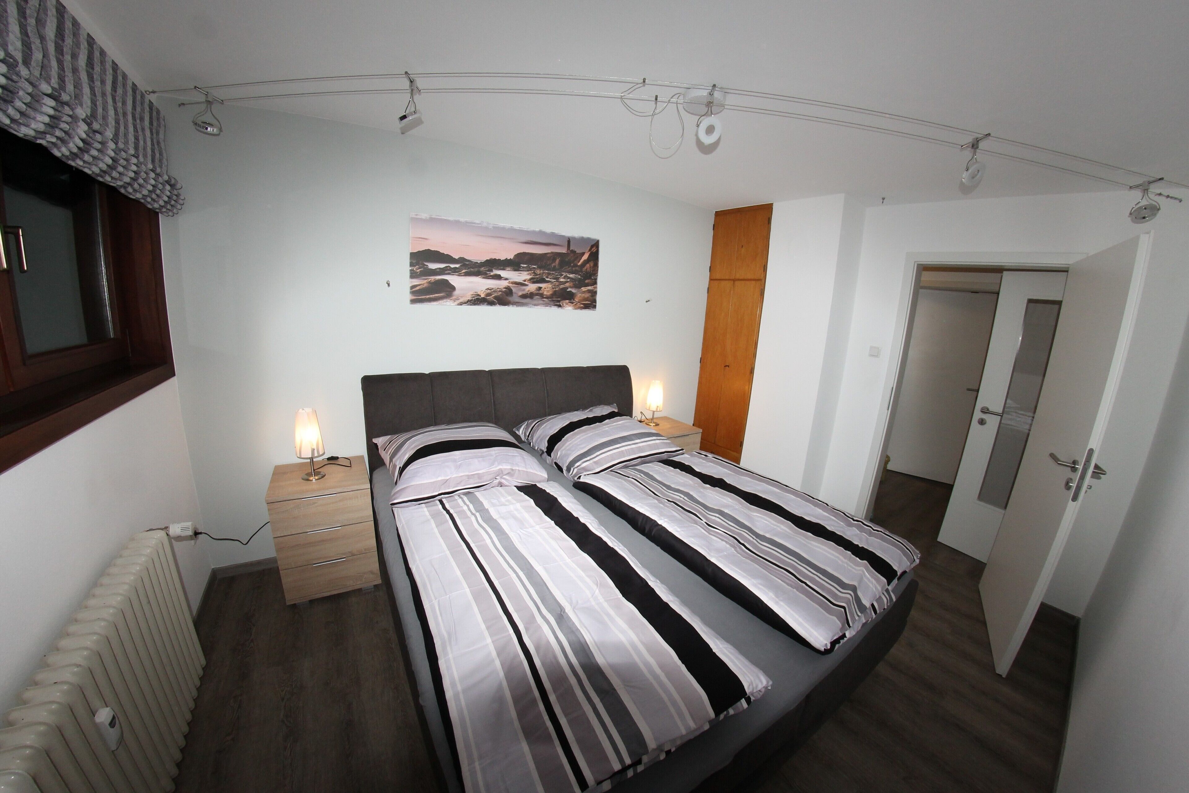 Ein Schlafzimmer mit Bett, Nachttischlampen, einem Holznachttisch, einem Heizkörper, einem Wandbild und einer Tür.