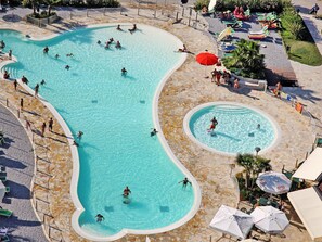 Une grande piscine extérieure dotée de plusieurs couloirs, entourée d’une terrasse en pierre et ombragée par des parasols.