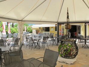 Un café en plein air avec des tables et des chaises sous une grande marquise, un bac à fleurs décoratif et un bâtiment en arrière-plan.