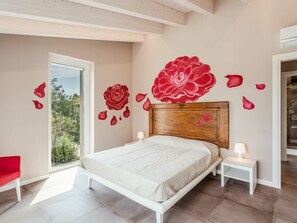 Une chambre avec un grand lit, une tête de lit en bois, une fenêtre donnant sur un paysage verdoyant et un motif floral rouge décorant un mur.