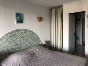 Une chambre avec une tête de lit recouverte de carreaux verts, un lit avec une couvre-lit à motifs, une lampe fixée au mur, un tableau et une porte donnant sur une autre pièce.