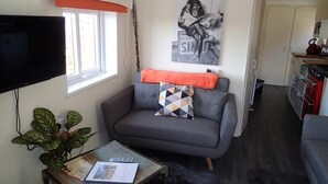 A compact living space with a grey sofa, a small table with a plant, a television, and a kitchen area in the background. - Rose Lodge, Eco Lodge with Hot Tub (Langport)