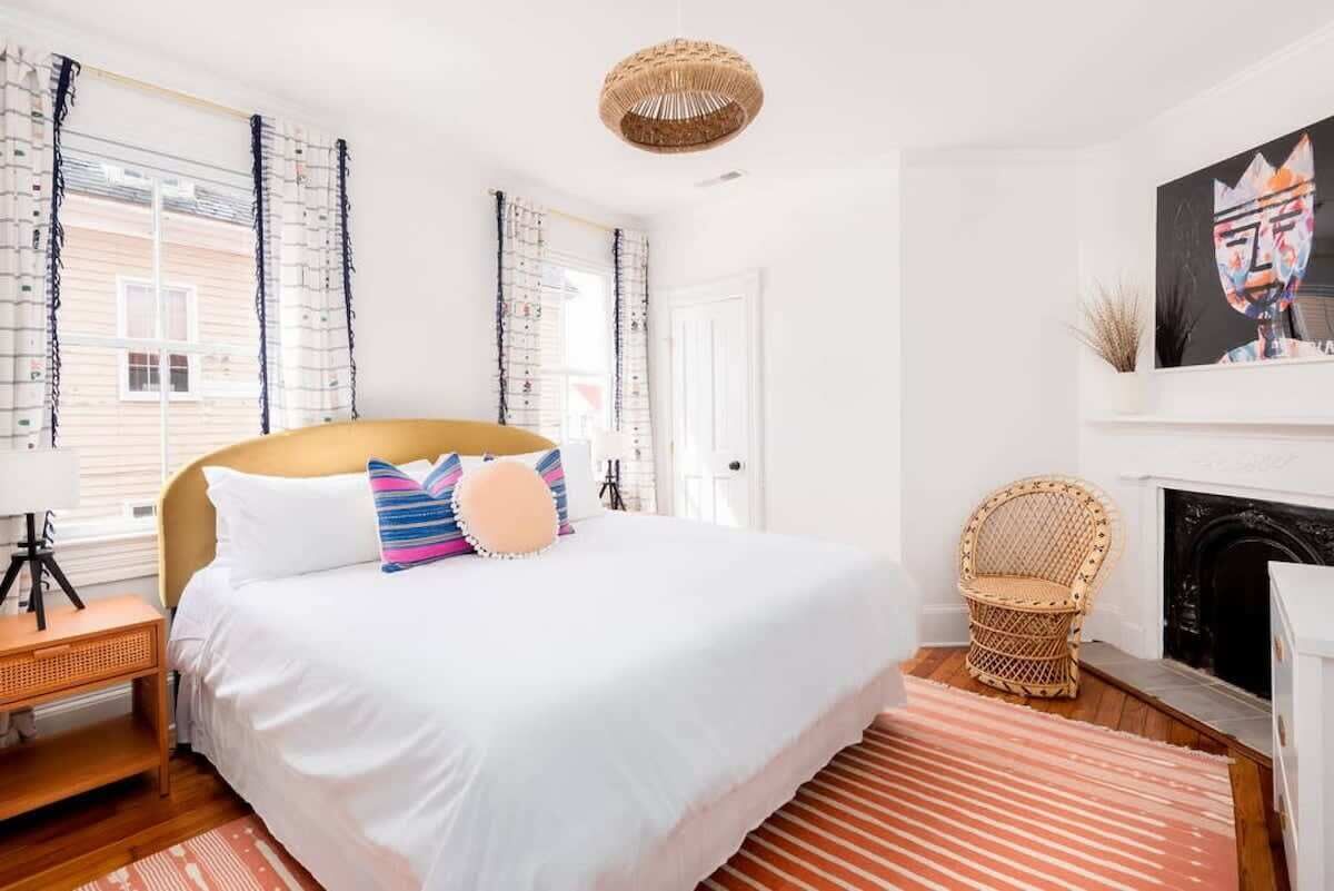 A bedroom with a large bed, a wicker chair, a fireplace, and a painting on the wall.