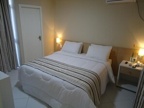 A neatly made bed with a striped blanket, two bedside lamps, and a nightstand with a phone and a vase of flowers. - Serra Plaza Hotel (Serrinha)