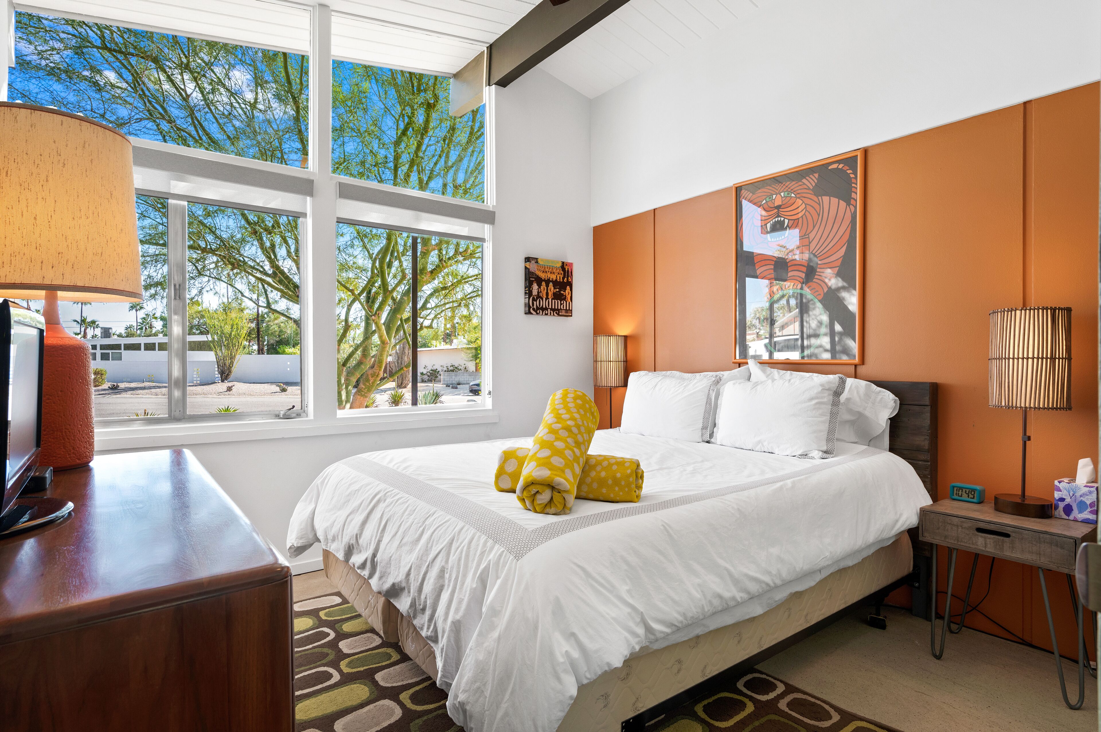 A hotel room with a large bed, a desk, a lamp, and a window with a view of trees and a parking lot.