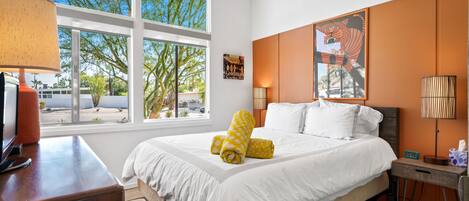 A hotel room with a large bed, a desk, a lamp, and a window with a view of trees and a parking lot.