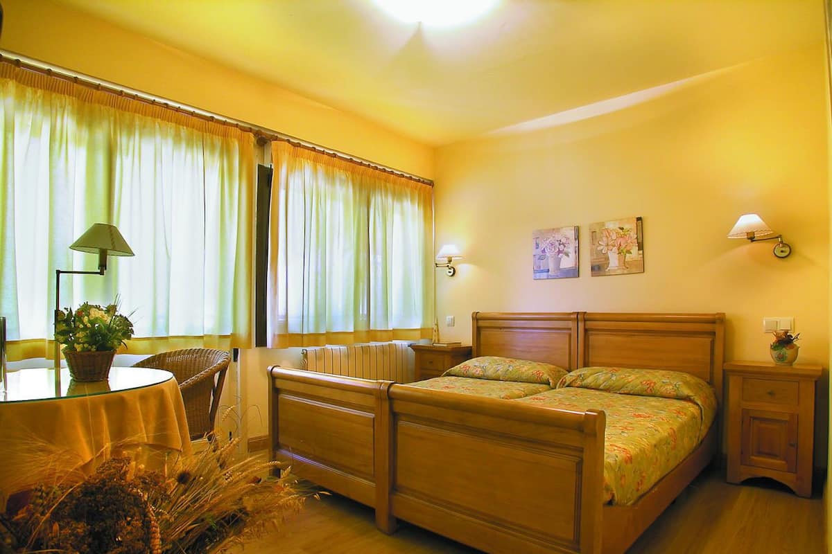 A bedroom with a wooden bed, a bedside table, a nightstand, a chair, a table with a lamp, and a window with curtains.