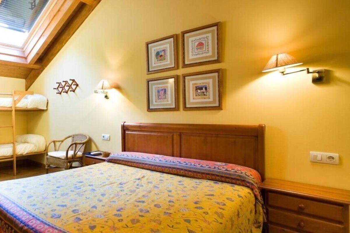 A bedroom with a bunk bed, a single bed, a wooden headboard, a bedside table, a chair, and a wall-mounted lamp.