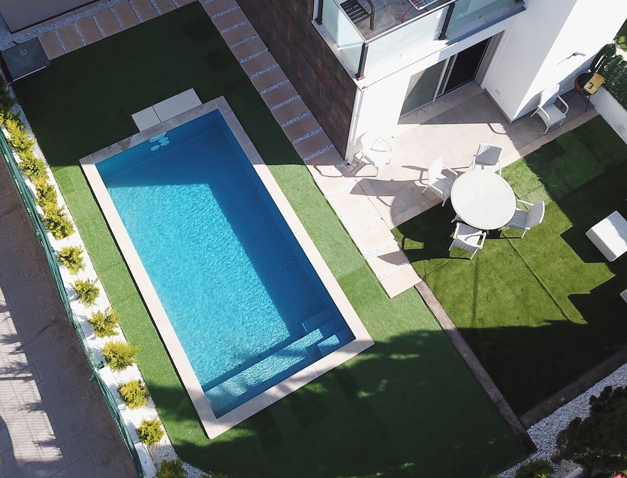 Una piscina rectangular rodeada de un césped cuidado, una zona de patio con mobiliario exterior y un edificio moderno con fachada de vidrio.