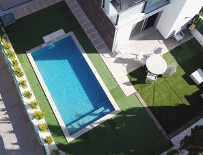A rectangular swimming pool surrounded by a well-maintained lawn, a patio area with outdoor furniture, and a modern building with a glass facade. - House with pool a few steps from the beach (Alcúdia)