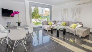 Ein modernes Wohnzimmer mit einem Glastisch, weißen Stühlen, einem grauen Sofa mit grünen Kissen und einem großen Fenster mit Blick ins Freie.