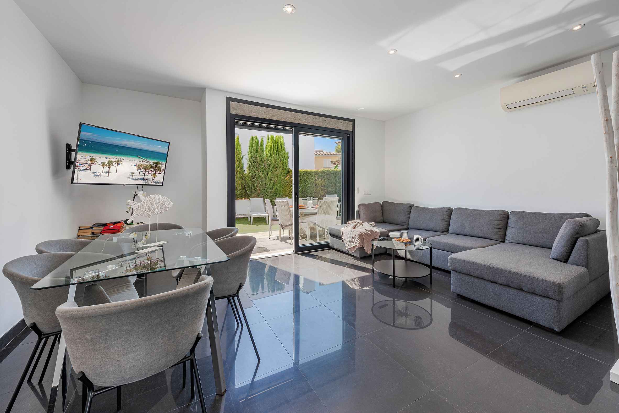 A modern living room with a grey sofa, a glass dining table with chairs, a flat-screen TV on the wall, and a sliding glass door leading to an outdoor area.