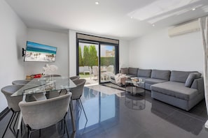 A modern living room with a grey sofa, a glass dining table with chairs, a flat-screen TV on the wall, and a sliding glass door leading to an outdoor area.