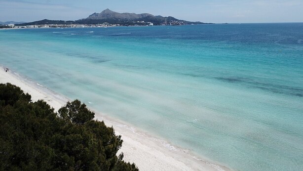 Ein Strand mit klarem türkisblauem Wasser, einem sandigen Ufer und einem gebirgigen Hintergrund.