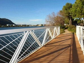 A wooden deck with white railings overlooking a body of water, with trees and buildings in the background. - House with pool a few steps from the beach (Alcúdia)