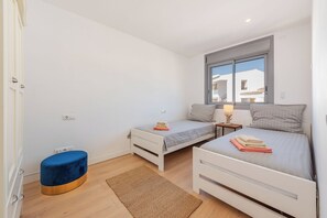 A bedroom with two beds, a small round ottoman, a rug, and a window with a view of buildings. - House with pool a few steps from the beach (Alcúdia)