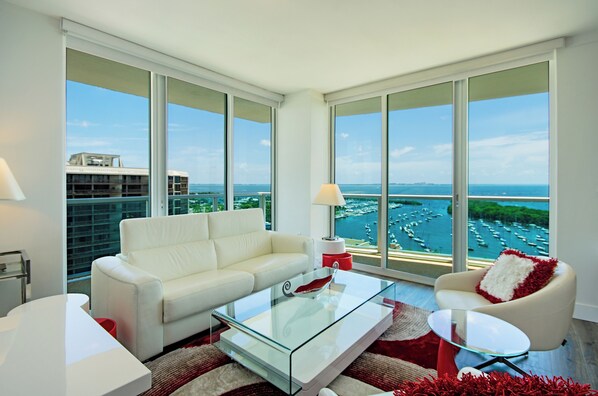 Un salón moderno con un sofá blanco, una mesa de centro de cristal y un sillón rojo. Amplias ventanas ofrecen vistas a un puerto deportivo con barcos.