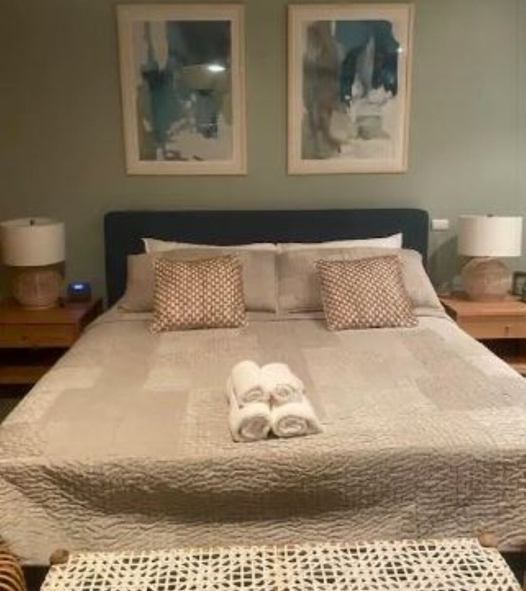 Ein ordentlich bezogenes Bett mit einem zusammengerollten Handtuch in der Mitte, zwei gemusterten Kissen und zwei gerahmte abstrakte Gemälde darüber.