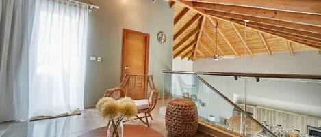 Une pièce spacieuse avec un plafond haut, un plafond en bois, une rambarde en verre, une table ronde, une chaise en osier et une porte.