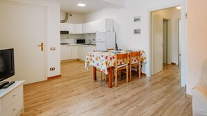 Een compacte keuken met witte kastjes, een magnetron en een koelkast. Er is een eettafel met stoelen en een tafelkleed met bloemenmotief.