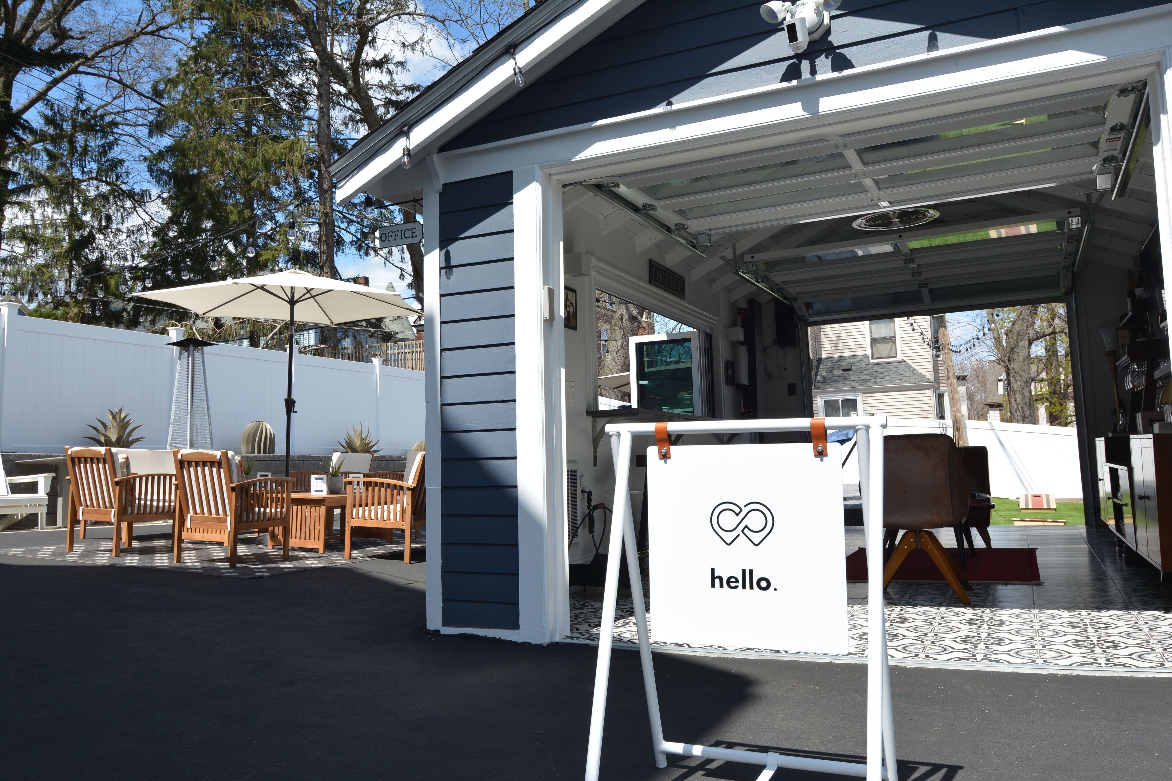Un espace extérieur aménagé avec des chaises et une table en bois, un patio couvert avec un parasol, et une pancarte sur laquelle on peut lire « bonjour ».