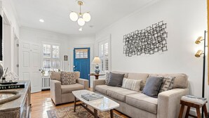 Ein Wohnzimmer mit einem beigen Sofa, zwei Sesseln, einem Couchtisch und einem gemusterten Teppich. Man sieht eine blaue Tür, ein Wandbild und eine Stehlampe.