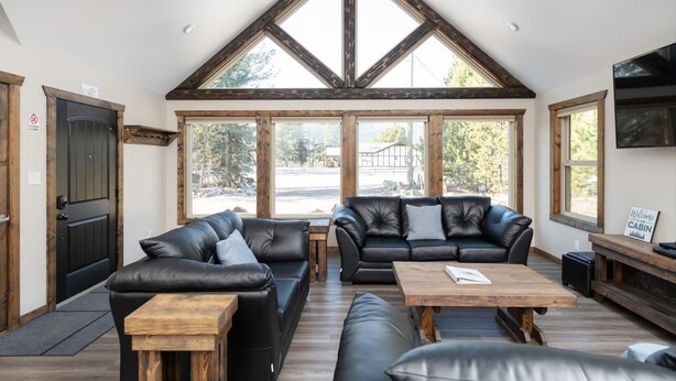 A spacious room with a high ceiling, large windows, a black leather sofa, a wooden coffee table, and a television on the wall. - Summit View Cabin Rental (British Columbia)