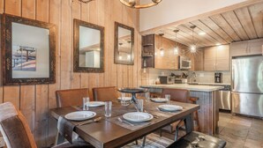Comedor con una mesa de madera preparada para cuatro, cocina con electrodomésticos de acero inoxidable y paneles de madera en las paredes.