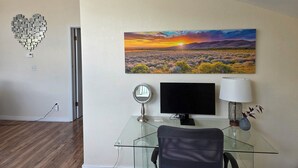 Une pièce avec un bureau en verre, un écran d’ordinateur, une lampe et un tableau représentant un paysage au coucher du soleil, accroché au mur.