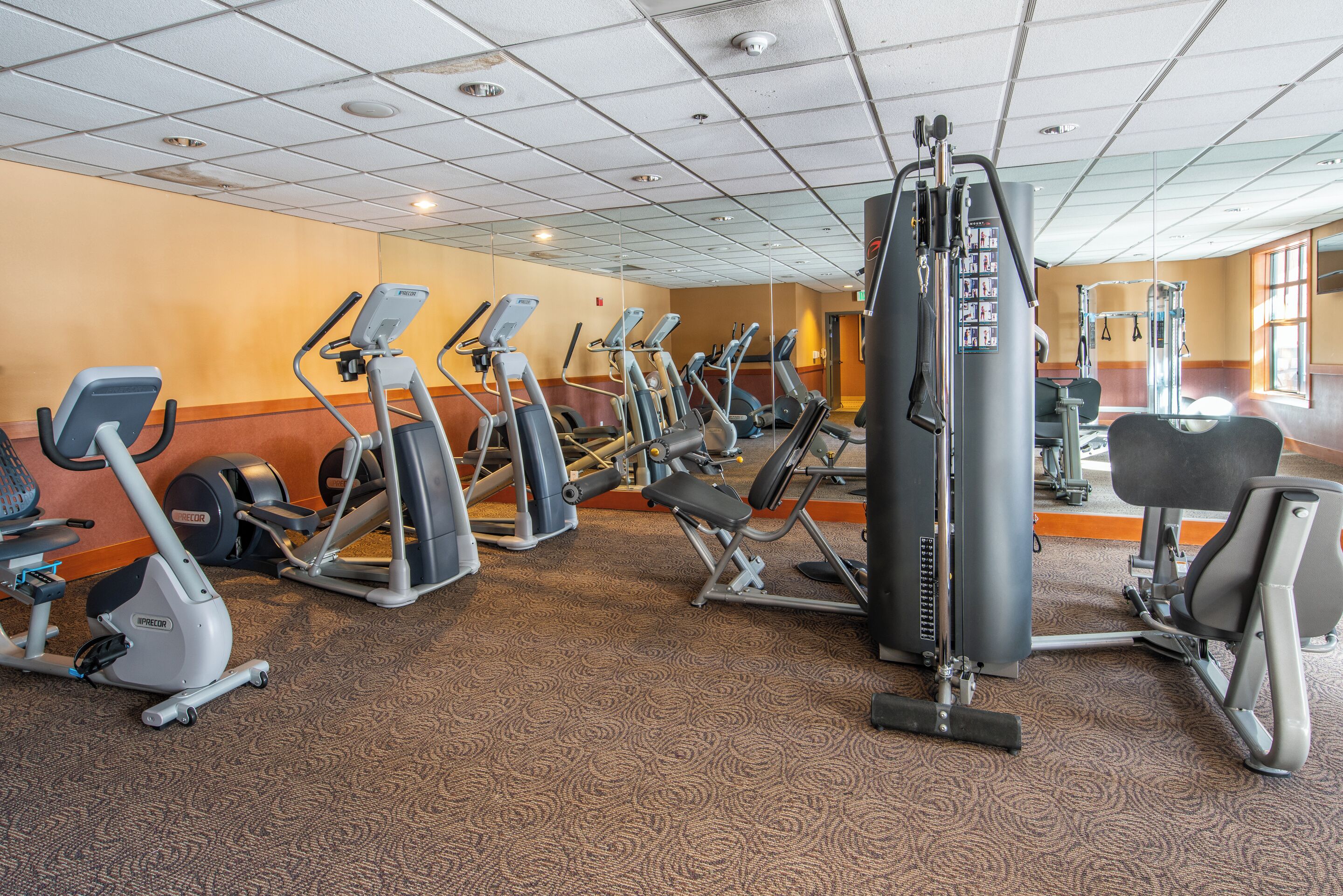 A gym with various cardio and strength training equipment, carpeted flooring, and a mirrored wall.