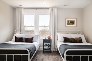 A bedroom with two beds, a nightstand with a lamp, a framed picture on the wall, and a view through a window. - Design-Forward East Nashville Townhome • 10 Min to Broadway | Rhett (Nashville)