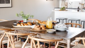 Un comedor con una mesa de madera dispuesta para el desayuno, que incluye bollería, frutas y un vaso de zumo de naranja.