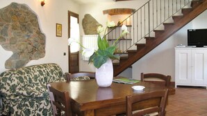 A dining area with a wooden table, chairs, a vase with flowers, a staircase, a sofa, and a television on a cabinet.