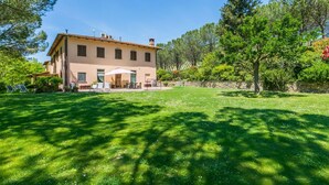 A two-story house with a patio, surrounded by a well-maintained lawn and trees. - Lovely apartment in villa for 4 people with WIFI, pool, TV, patio, pets allowed and panoramic view (Montespertoli)