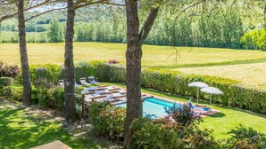 Un espace piscine doté de chaises longues et de parasols, entouré d’arbres et d’un champ d’herbe.