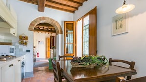 Uma cozinha com piso de tijolos, vigas de madeira no teto, uma mesa de jantar com cadeiras e uma janela com cortinas.