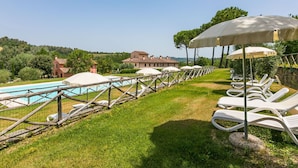 Uma área de piscina com espreguiçadeiras e guarda-sóis, uma piscina e um edifício ao fundo.