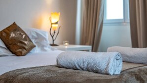 A neatly made bed with a white blanket and a brown pillow, a white towel on the bed, a bedside table with a candle, and a window with curtains. - Bright Airy Spacious Property (Passy)