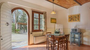 Uma sala de jantar com mesa e cadeiras de madeira, janela com vista para árvores, ventilador de teto e micro-ondas sobre uma pequena mesa.