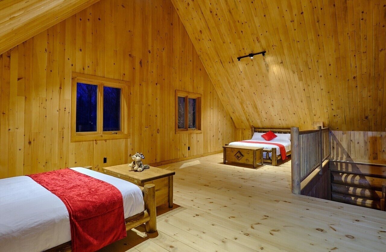 A wooden cabin with two bedrooms, each with a bed and a window. There is a small table with a teddy bear in the first room.