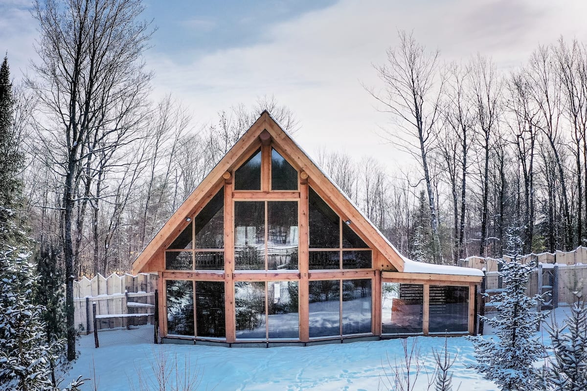 Una cabaña de madera con ventanas grandes, rodeada de árboles cubiertos de nieve y un cielo despejado.