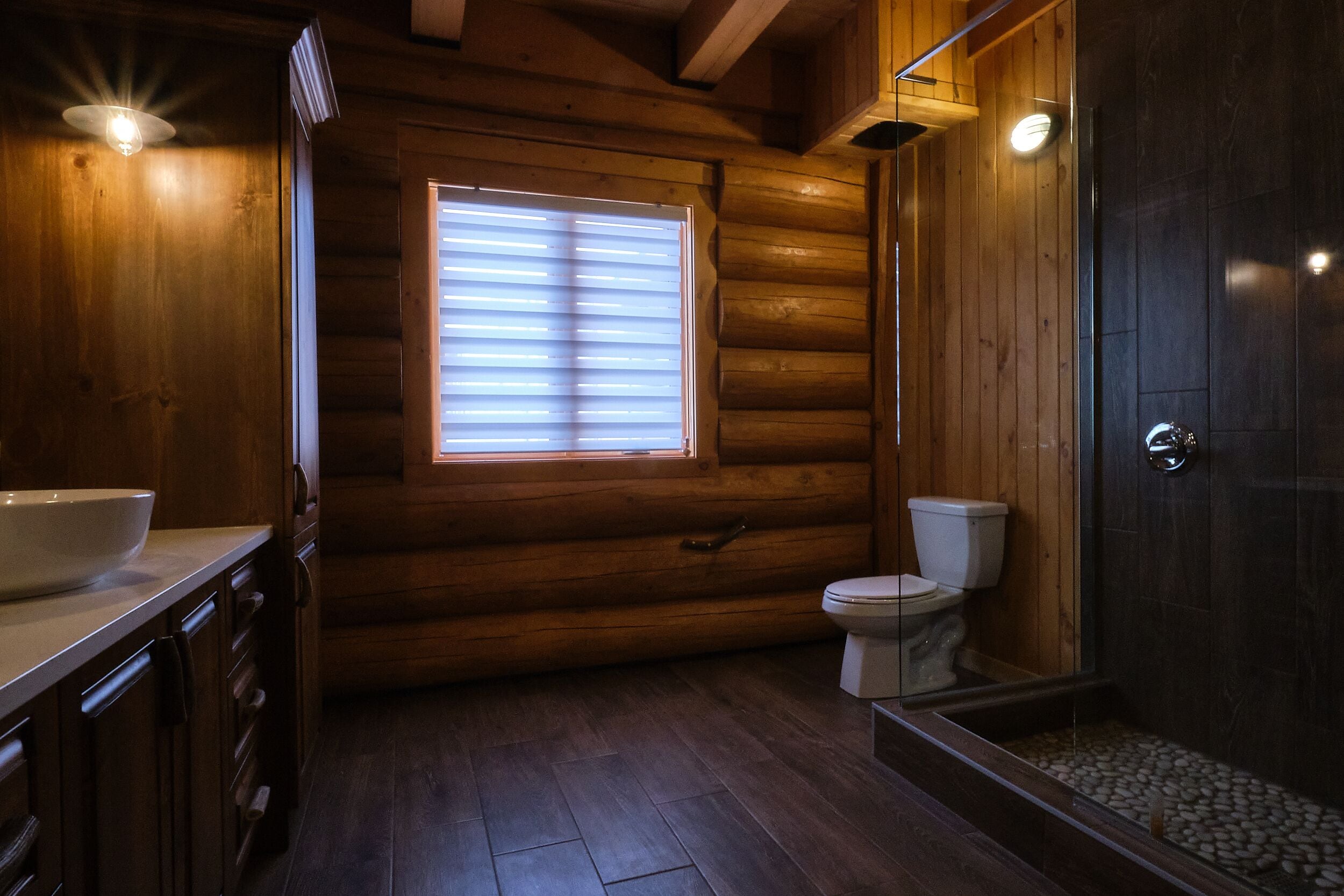 Une salle de bain rustique, aménagée dans une cabane en rondins, avec un intérieur en bois, des toilettes blanches, une douche en verre et une grande fenêtre.