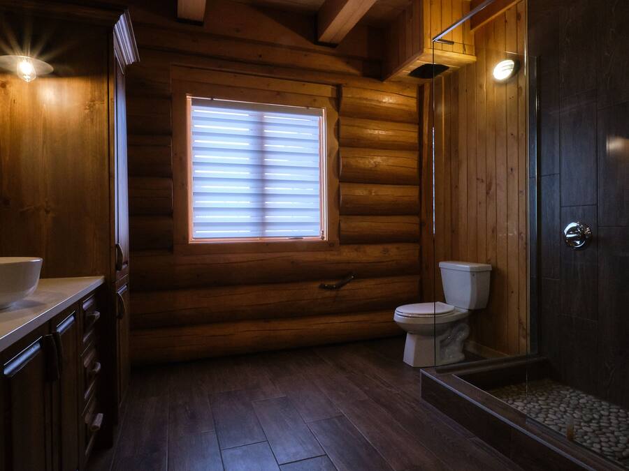 A log cabin bathroom with a wooden interior, a white toilet, a glass-enclosed shower, and a large window.