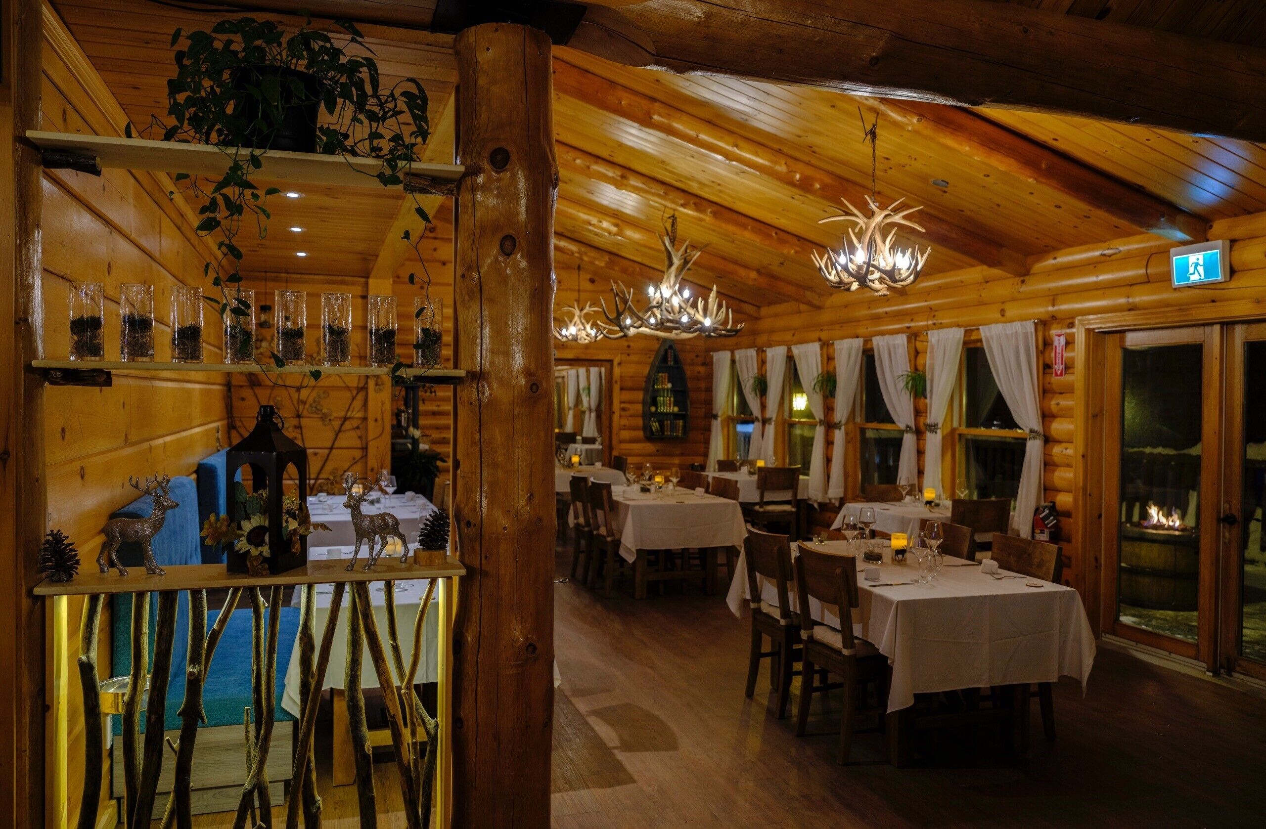 Un intérieur chaleureux, avec des murs en bois, des tables dressées pour le repas, un lustre et une cheminée visible à travers des portes vitrées.