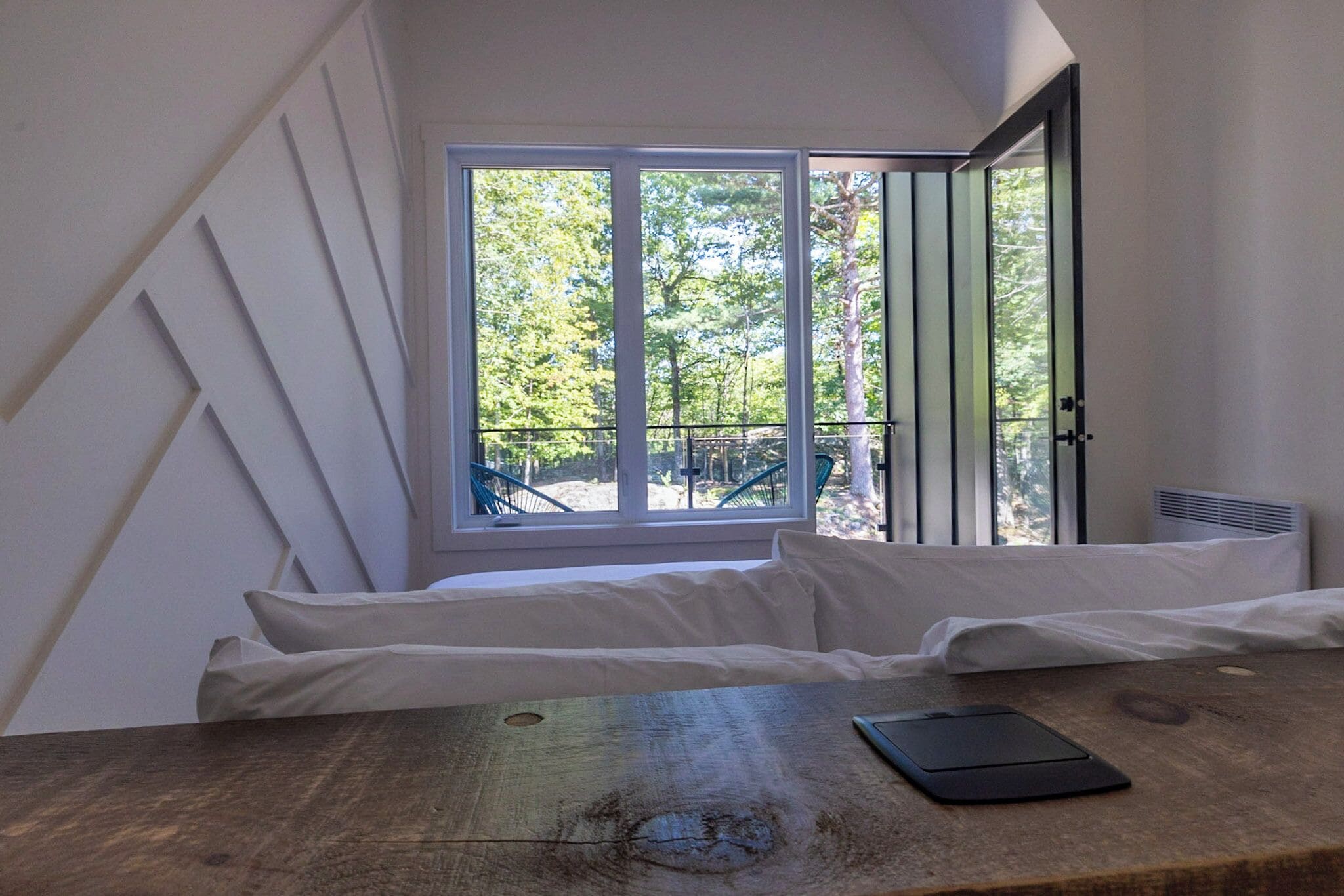 A bedroom with a large window, a wooden table, and a bed with white bedding.