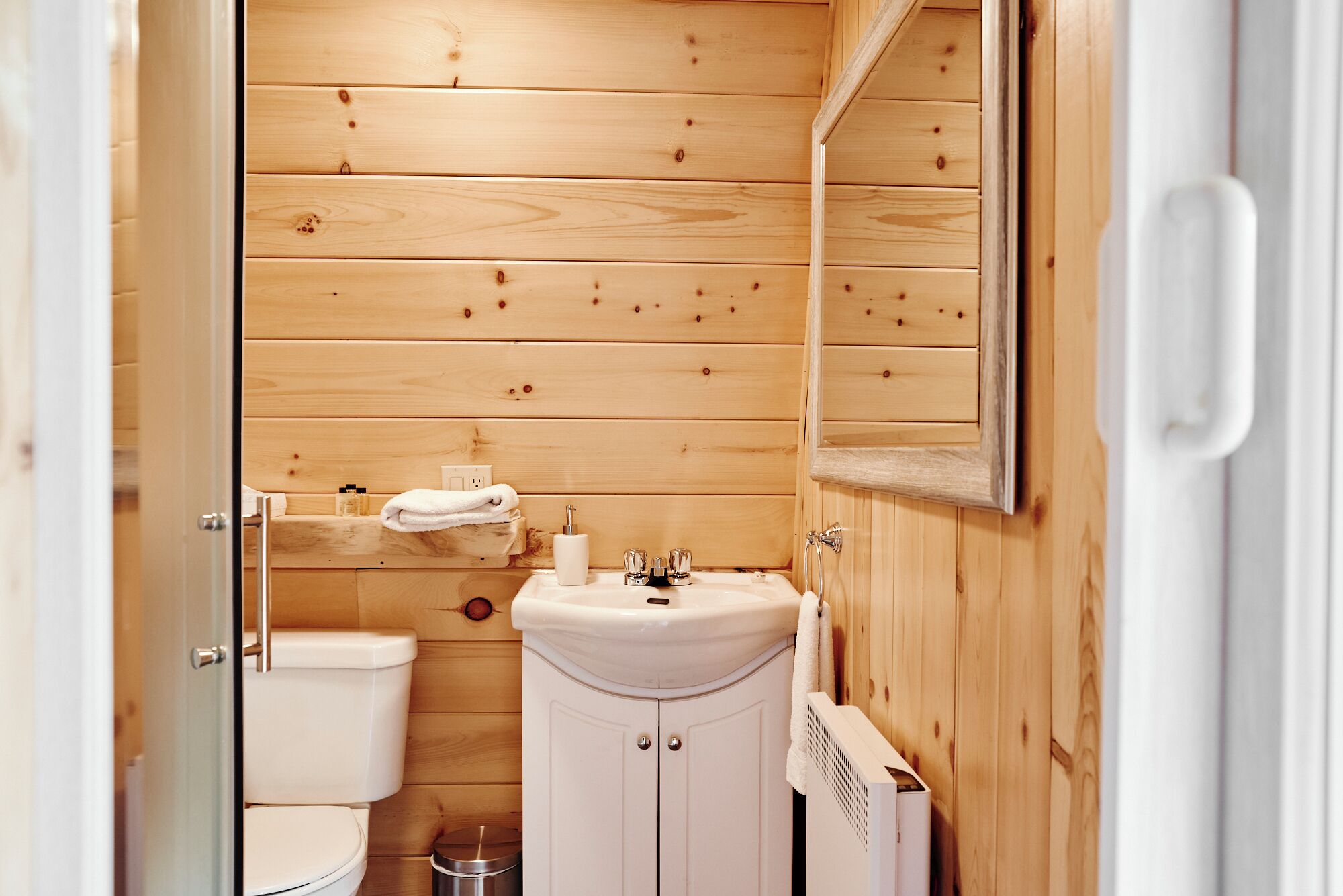 Ein kleines, kompaktes Badezimmer mit Holzwandverkleidung, einer weißen Toilette, einem Waschbecken mit Spiegel und einem Handtuchhalter.