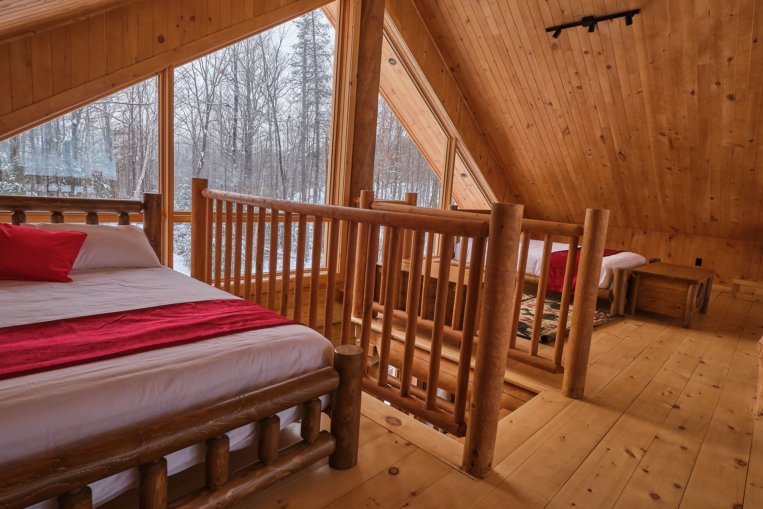 A wooden cabin bedroom with a bed, a red pillow, and a view of a snowy forest through large windows.