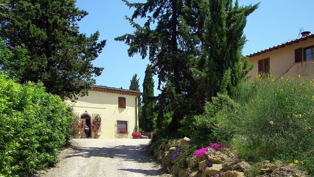 Un edificio rustico con un vialetto sterrato che conduce a una porta di legno, immerso nel verde e circondato da piante fiorite.