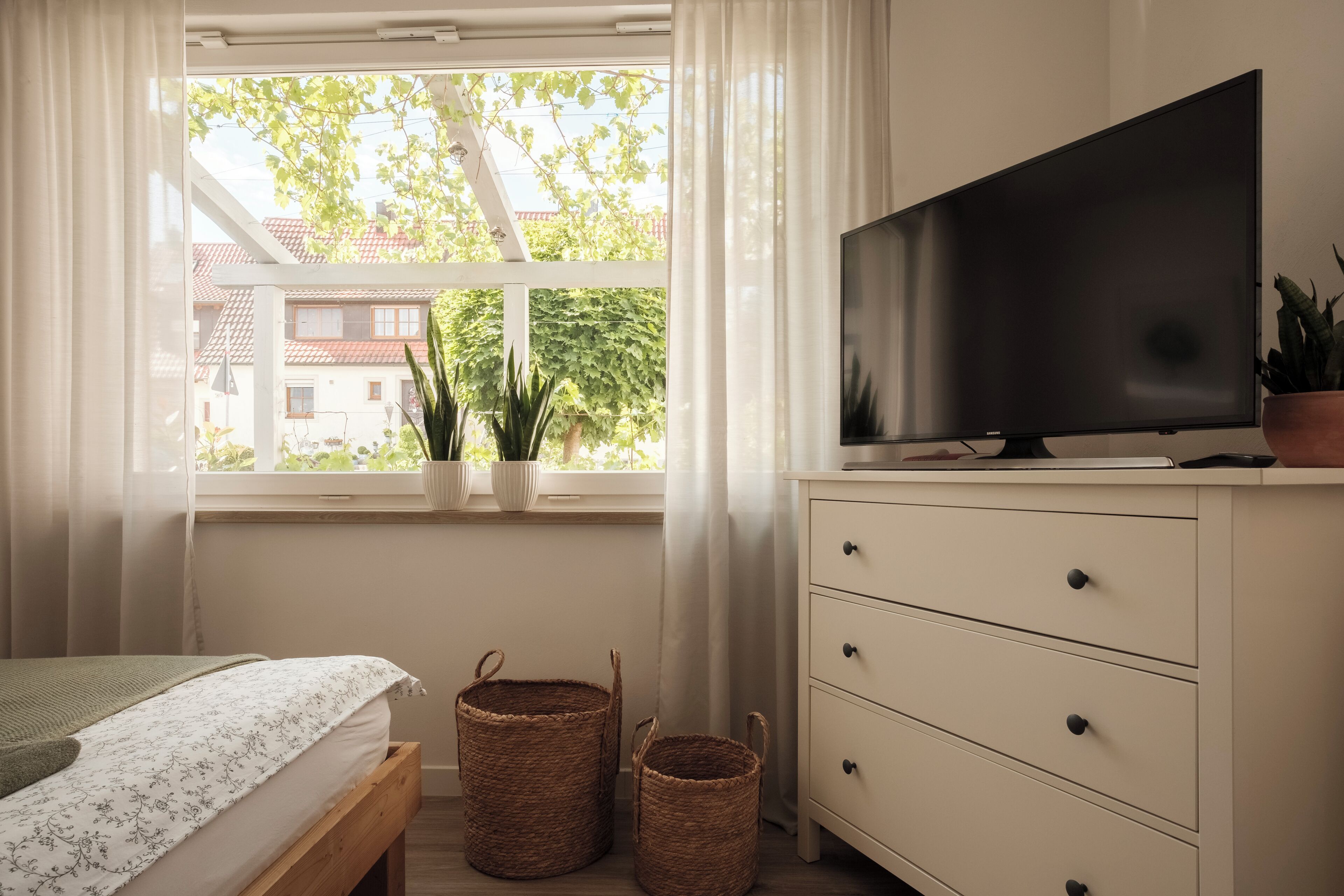 Une chambre à coucher avec un lit, une télévision, une fenêtre avec des rideaux et deux paniers en osier.