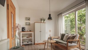 Un salon chaleureux avec un canapé en bois, un meuble blanc, une petite table sur laquelle est posé un vase, et une grande fenêtre donnant sur un paysage verdoyant.
